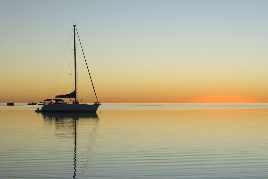 En sejlbåd for anker på roligt vand ved solnedgang. Himlen er orange og gul, og vandet reflekterer de gyldne farver.